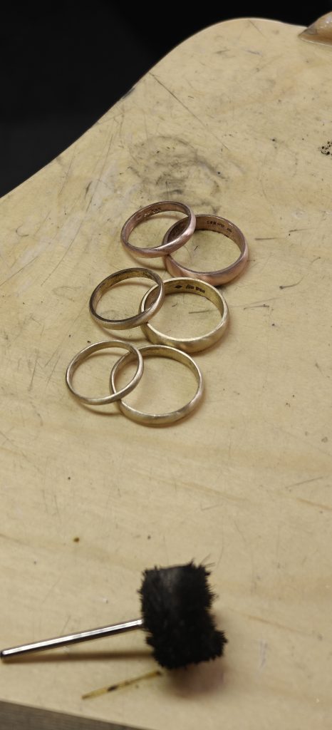 Mehrere Eheringe aus verschiedenen Metallen (Gold, Silber, eventuell Roségold) liegen auf einer hölzernen Werkbank. Unten im Bild befindet sich ein schwarzer Polieraufsatz mit Metallschaft, der zum Bearbeiten von Schmuckstücken verwendet wird.
